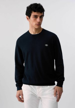 Młody mężczyzna ubrany w czarną koszulę z długim rękawem z małym logo na piersi oraz białe spodnie, na tle jednolitego, jasnego tła.