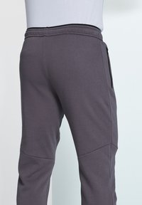 Grey sweatpants feature a smooth texture, elastic waistband, and a side zip pocket. Enhanced with stitched panel details for added shape.