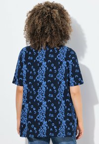 Námořnická modrá tričko s krátkými rukávy, které má opakovaný vzor světle modrého textu v různých orientacích. Měkká, texturovaná tkanina.