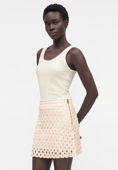 Γυναίκα που φοράει ένα αμάνικο εκρού τοπ και μια μπεζ φούστα από δαντέλα με χρυσό φερμουάρ, στέκεται μπροστά από λευκό φόντο.