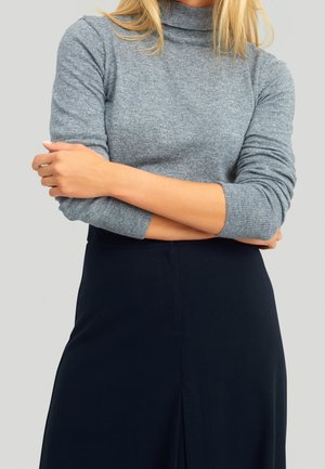 Vrouw met gevouwen armen, gekleed in een grijze coltrui met lange mouwen en een zwarte rok, tegen een effen lichte achtergrond.