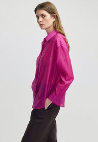 Dlouhý rukáv, magentová button-up košile, vyrobená z průsvitného materiálu, s špičatým límcem a volným střihem, spárovaná s tmavými kalhotami.
