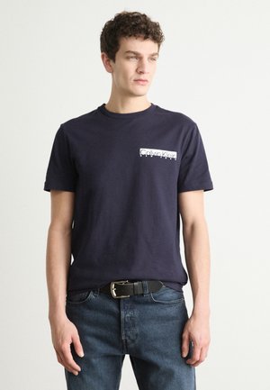 Tricou din bumbac navy cu mâneci scurte, având un logo alb Calvin Klein în zona stângă a pieptului, asociat cu blugi denim albaștri și un centură neagră.