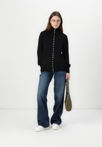Maglione nero con colletto alto e fila centrale di bottoni rotondi. Abbinato a jeans a gamba larga e sneakers bianche. Tiene in mano una piccola borsa verde.
