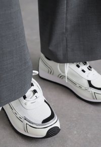 Baskets de sport blanches avec des accents noirs et gris, empeigne en mesh, semelle rembourrée et semelle extérieure en caoutchouc noir ; associées à un pantalon gris.