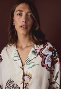Woman with wavy brown hair wearing silver hoop earrings and a cream blouse with colorful floral and zebra prints, against dark brown background.