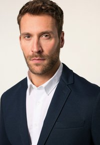 Blazer blu navy con tessuto texturizzato, taschino sul petto, abbinato a una camicia bianca con colletto. Vista ravvicinata, con focus su parte superiore del corpo e testa.