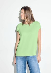 Blusa verde chiaro con maniche corte, scollatura rotonda e taglio dritto. Abbinata a jeans blu, esposta su uno sfondo bianco.