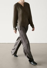 Blouse à manches longues vert olive avec patte de boutonnage et poignets ; associée à un pantalon large gris foncé, à la coupe fluide. Chaussures noires.