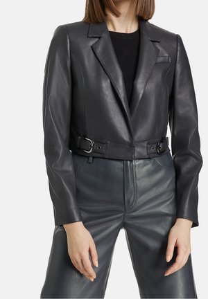 Femme portant un blazer en cuir noir ajusté avec des détails à boucle et un pantalon en cuir noir assorti, debout les mains détendues.