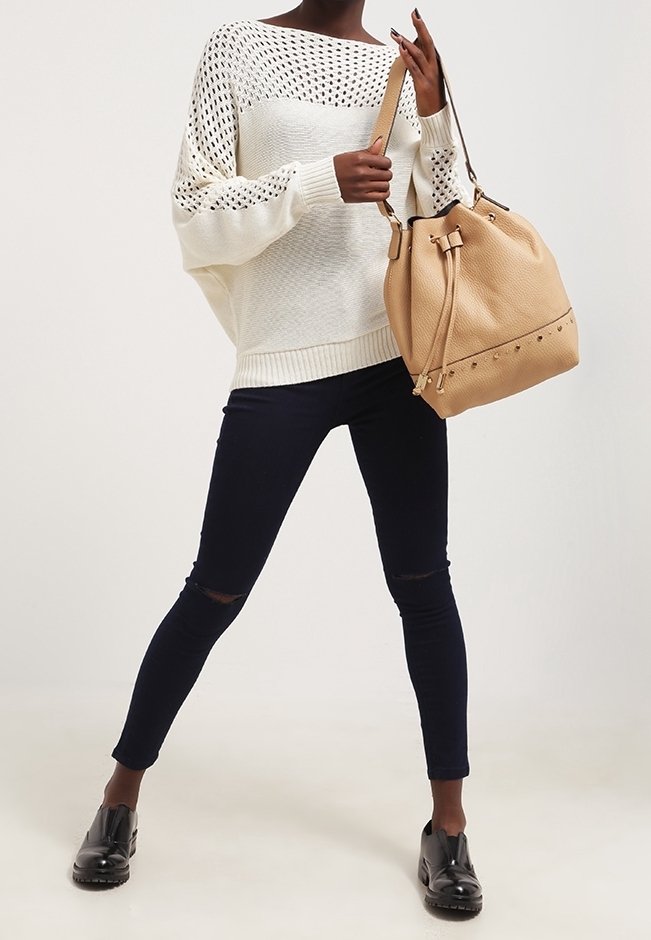 Femme portant un pull en maille blanc, un jean skinny noir déchiré, des chaussures noires, tenant un grand sac seau en cuir marron clair par sa sangle.