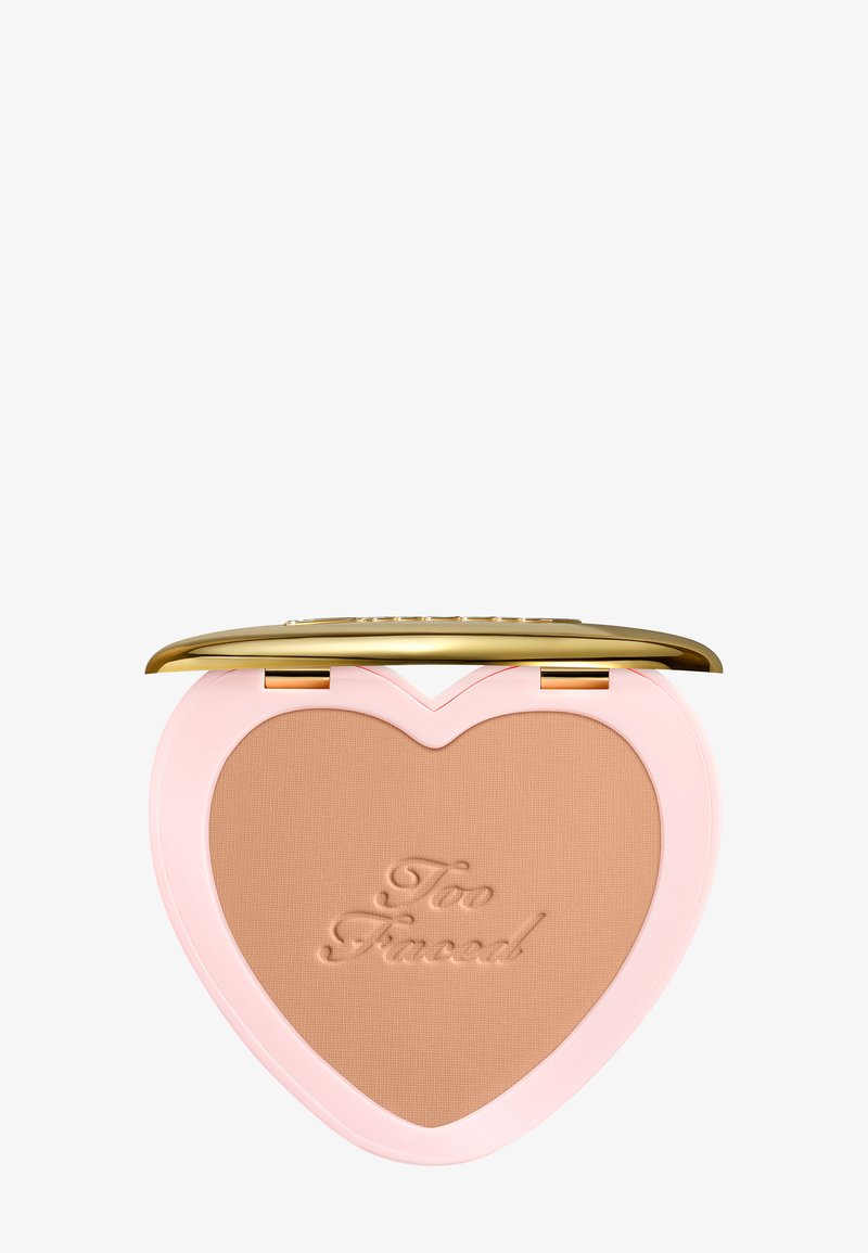 Poudre bronzante compacte en forme de cœur avec le logo "Too Faced" en relief, boîtier rose et couvercle à charnière dorée, ouvert sur fond blanc.