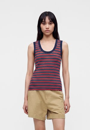 Ung kvinde iført en ærmeløs top med navy, rød og hvid striber og beige shorts, stående foran en ensformet lys grå baggrund.