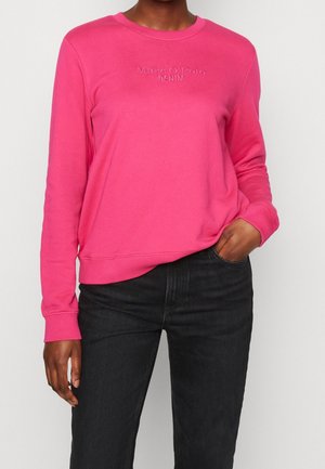 Person iført en lyserød langærmet sweatshirt med teksten "Marc O’Polo Denim" og sorte jeans, hånd i lommen, står foran en ensfarvet baggrund.