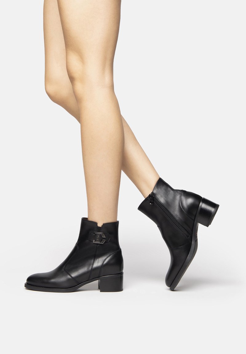 Schwarze Leder-Stiefeletten mit einem Blockabsatz, seitlichem Reißverschluss und einer dekorativen Metall-Schnalle an der Vorderseite. Glatte Textur und polierte Oberfläche.
