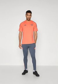 Koralové sportovní tričko s černými doplňky a logem Adidas, v kombinaci s přiléhavými šedými tepláky s korálovými bočními pruhy a černými teniskami.