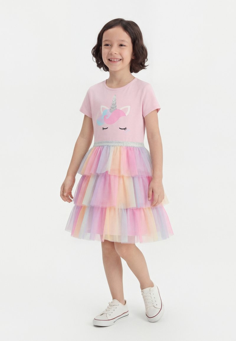 Κορίτσι που χαμογελά, φοράει ροζ φόρεμα με κοντά μανίκια και στάμπα μονόκερου, πολύστιλη παστέλ φούστα από τούλι και λευκά αθλητικά παπούτσια, στέκεται σε λευκό φόντο.