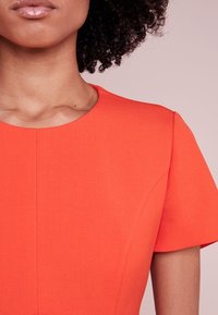 Robe orange à encolure ronde et manches courtes. Le tissu est texturé avec des détails de coutures visibles et une silhouette ajustée.