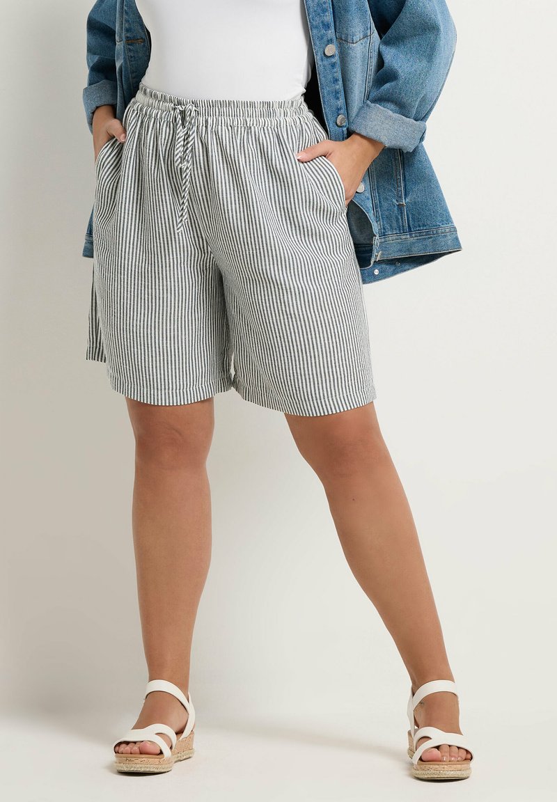 Persona che indossa pantaloncini a righe con coulisse, sandali bianchi, top bianco e giacca di jeans blu con le mani nelle tasche, in piedi contro uno sfondo semplice.