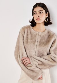 Giacca corta in finta pelliccia di un morbido beige, con scollo rotondo, maniche lunghe con ampi polsini e una texture liscia.