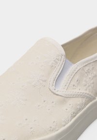 Κρεμ παπούτσι τύπου slip-on με λευκή κεντημένη λουλουδένια διακόσμηση και ελαστικές πλευρικές επιφάνειες, με λευκή λαστιχένια σόλα.