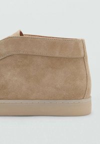 Beige mockaskinnsko med en slät yta, låg profil och en sydd söm längs sidan. Har en ljusgummisula.