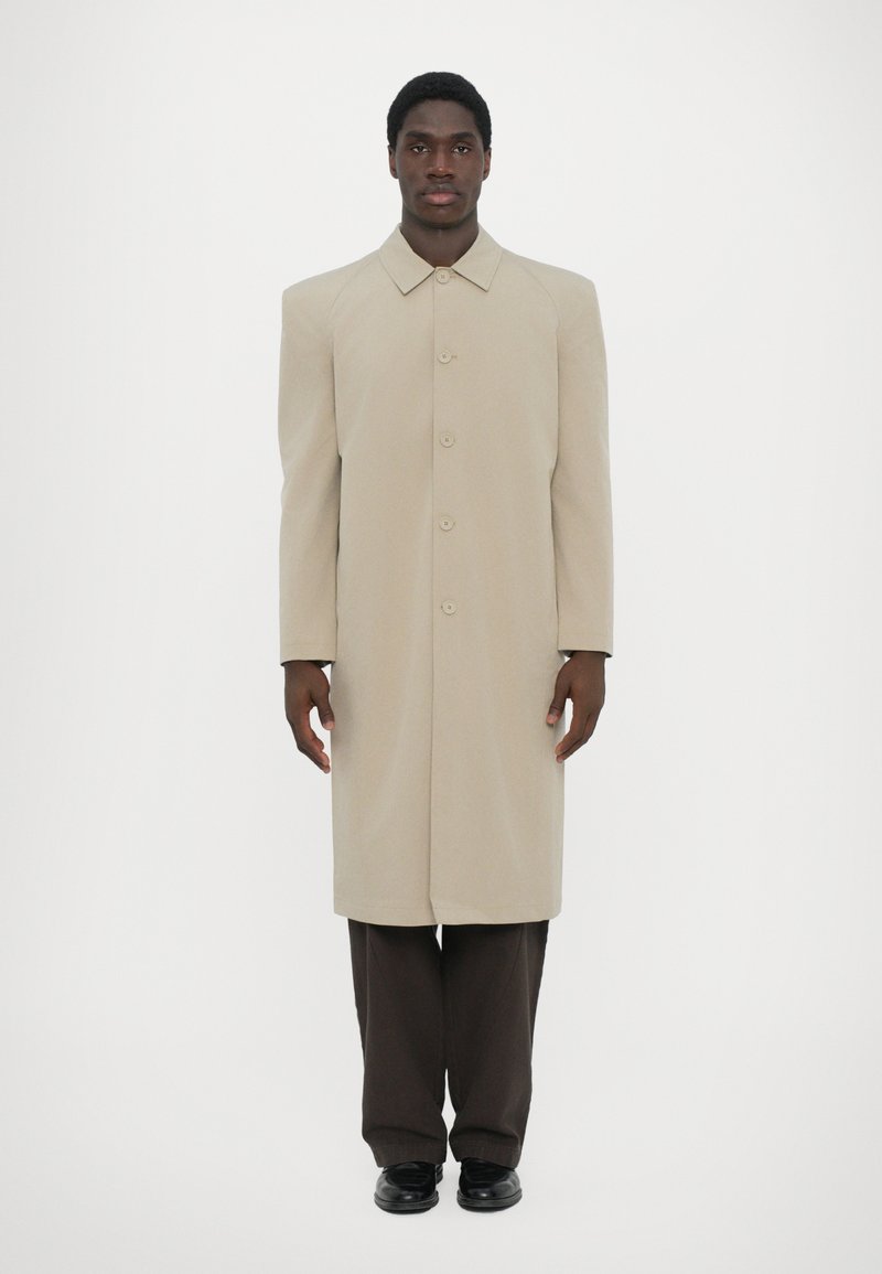 Beige long coat made of textured fabric with a straight cut, large collar, and button front; paired with dark brown trousers and black shoes.