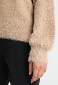 Beige pluizige trui met ribbelstructuur en wijde mouwen, gemaakt van zacht, fluffy materiaal. Hand rustend nabij de zoom.