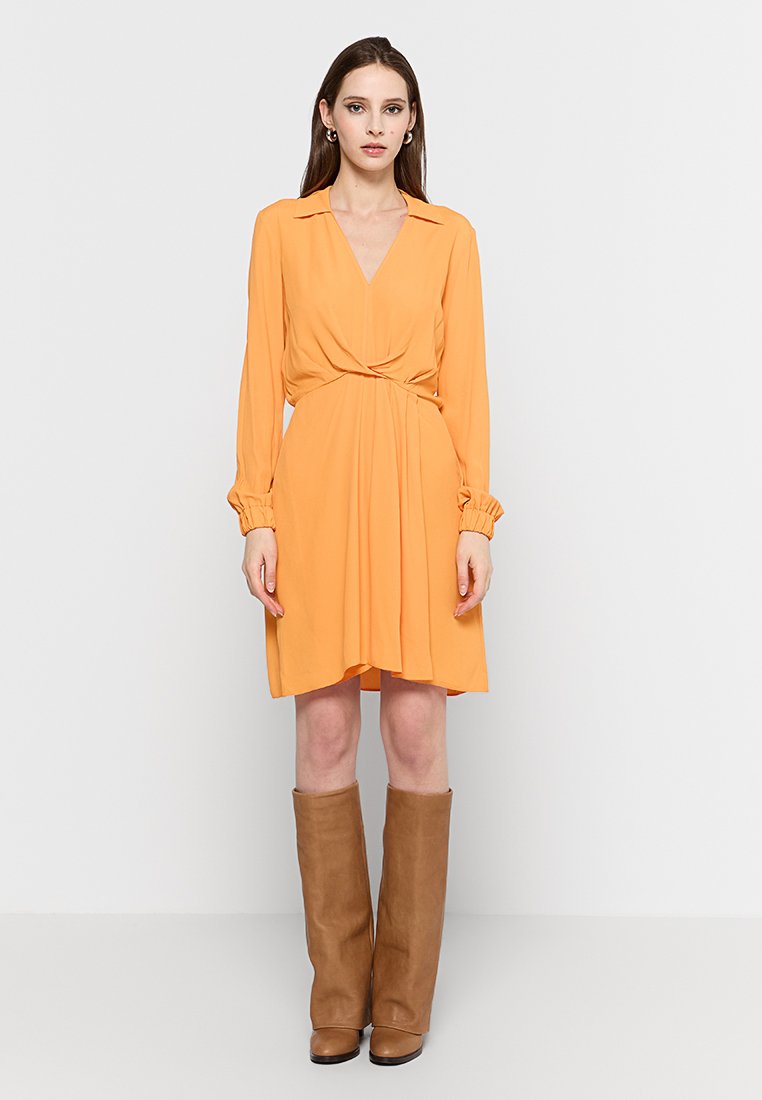 Pomarańczowa sukienka w wzór kopertowy z długimi rękawami, kołnierzykiem i marszczonymi mankietami. Siluetka w kształcie litery A z wiązanym w talii pasem, zestawiona z wysokimi brązowymi botkami.