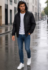 Homme marchant dans une rue mouillée de la ville, portant une veste de pluie noire à capuche, un t-shirt blanc, un jean bleu et des baskets blanches par une journée pluvieuse.