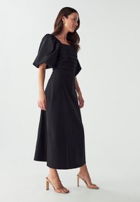 Black dress with puffed sleeves, square neckline, and horizontal pleats at the bodice. Floor-length with a straight silhouette and side slit.