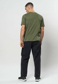 Mann i olivengrønn t-skjorte, svarte jeans og svarte joggesko står og vender bort fra et enkelt lyst bakgrunn.