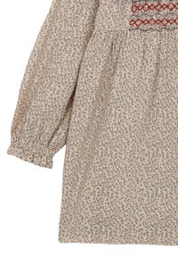 Bluzka w kremowym kolorze z motywem kwiatowym, wykonana z lekkiej tkaniny, z rozszerzanymi rękawami i marszczeniami. Na piersi znajduje się czerwona haftowana dekoracja.