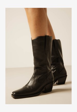 Čierne kožené kovbojské čižmy do polovice lýtka so špicatými špičkami a blokovým podpätkom, nosené na bosých nohách na neutrálnej pozadí.