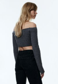 Grauer schulterfreier Cropped-Pullover mit gerippter Textur, langen Ärmeln und Bauchfreiheit, kombiniert mit hochgeschnittenen schwarzen Jeans.