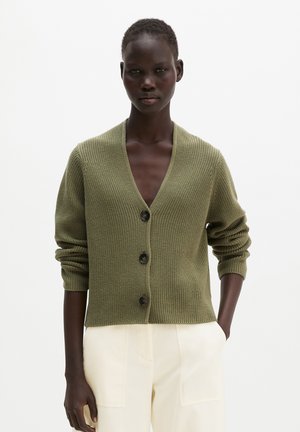Femme portant un cardigan tricoté vert olive à col en V, boutonné, manches longues et pantalon crème, debout avec une main dans la poche.