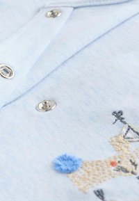 Chemise bleu clair avec une texture lisse, dotée de boutons-pression en argent et d'un motif brodé incluant des accents bleu et beige.