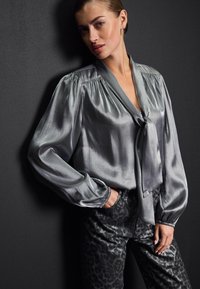 Bluză gri mătăsoasă cu guler legat, mâneci bufante și detalii plisate, asociată cu pantaloni negri cu imprimeu leopard. Texturată și netedă.