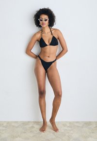 Svart bikini med trekantstopp og lavt liv, laget av strukturert stoff. Brukt med hvite katteøyne-solbriller, står på en steingulv.