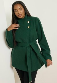 Vihreä villasekoitekotöö, jossa on vyötärölle sidottu vyö, kaksoisnapitus kultasävyisillä napilla ja muotoillut hihat.