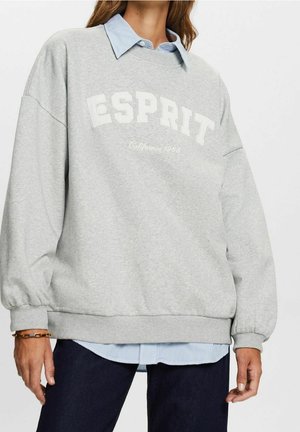 Person trägt ein hellgraues Esprit-Sweatshirt über einem blau gestreiften Hemd, kombiniert mit dunklen Jeans und einem goldenen Armband am linken Handgelenk.