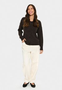 Eine Frau mit langen braunen Haaren trägt einen dunkelbraunen, knöpfbaren Pullover, cremefarbene weite Hosen und schwarze Ballerinas. Sie lächelt mit einer Hand in der Tasche.