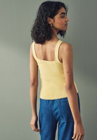 Canotta gialla a coste con schiena quadrata e orlo scallop, abbinata a jeans di denim blu. La modella ha i capelli ricci ed è posata di profilo.