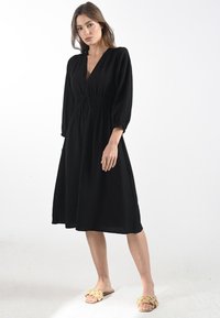 Abito nero al ginocchio con scollatura a V profonda, vita elasticizzata e maniche a sbuffo. Tessuto strutturato con una vestibilità rilassata e una silhouette fluida.