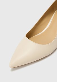 Gros plan sur une chaussure plate beige à bout pointu avec une tige en cuir lisse et une semelle intérieure couleur tan sur un fond clair.