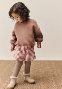 Felpa marrone, pantaloni shorts in gingham rosa, calze marroni testurizzate, stivaletti beige con pelliccia. Bambino in piedi su un tappeto decorato, guarda verso il basso.