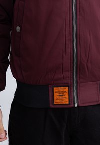 Mahoniekleurig vliegjack met zwarte geribde zoom, zilveren rits, zijvak met drukknop en oranje label met de tekst "Bombers Original" aan de voorkant.