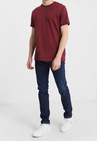 Vinröd t-shirt med kort ärm och rund halsringning. Buren med mörkblå slim-fit jeans och vita sneakers, mot en enfärgad bakgrund.