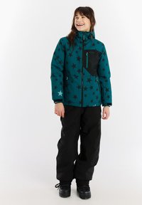 Zimska jakna u tirkiznoj boji s crnim uzorkom zvijezda, s prednjim zatvaračem i džepovima, upairana s crnim snježnim hlačama i tamnim čizmama.