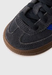 Detailný záber na špičku čiernej semišovej tenisky s textúrovanou gumovou podrážkou, čiernymi šnúrkami a modrým cikcakovým akcentom na boku.